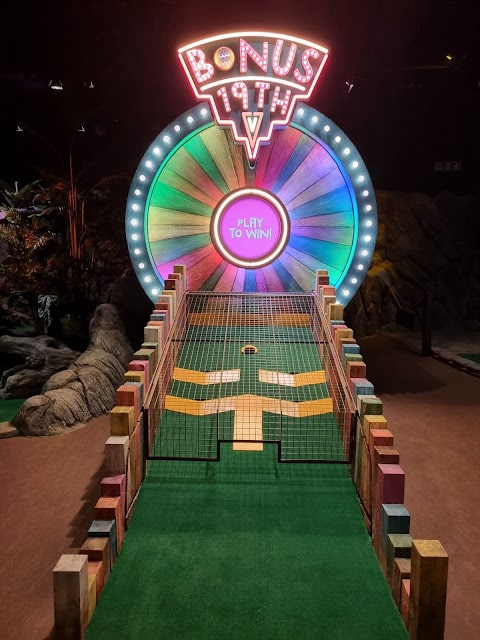 A brightly lit mini-golf hole features a colourful, illuminated wheel labelled "BONUS 19TH" and "PLAY TO WIN!" at the end. The course has a green surface, wooden borders, and raised barriers leading up to the spinning wheel backdrop.