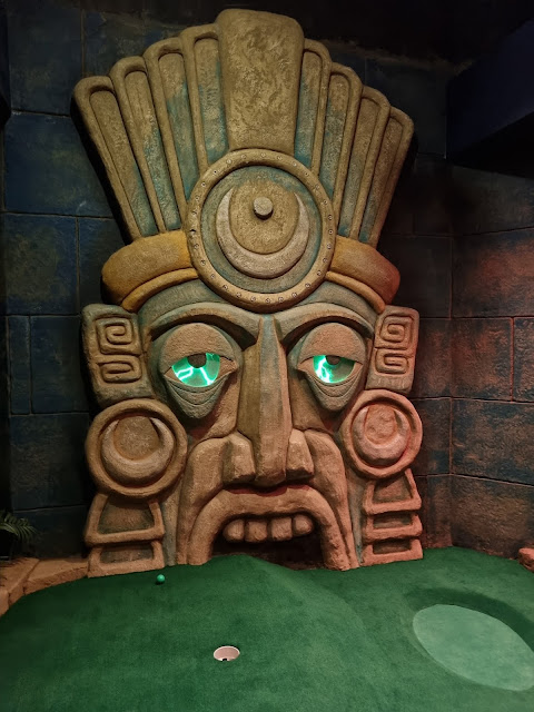 A large stone sculpture of an Aztec-style face with glowing green eyes stands behind a green mini-golf putting area. The face has a headdress, geometric designs, and a mouth opening leading to a hole on the putting green.