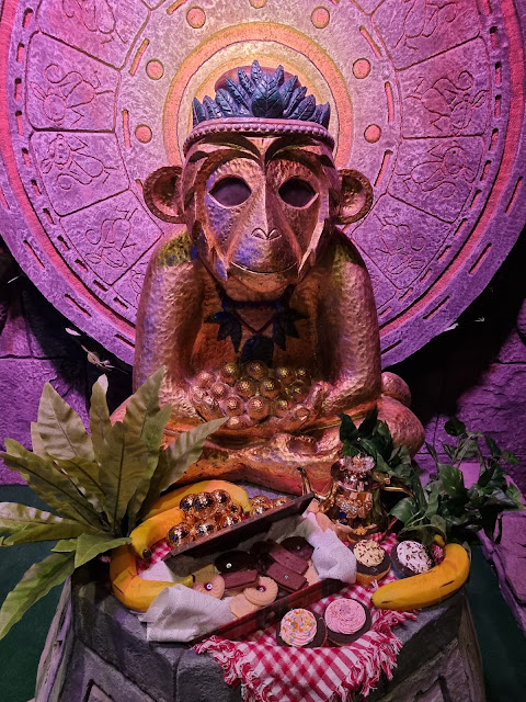 A gold monkey statuette with a beaded necklace sits cross-legged in front of a decorative, circular stone backdrop. In front, there’s a tray of assorted biscuits, chocolates, and leaves, set on a yellow cloth atop a stone plinth, under pink lighting.