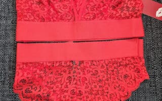 A red lace bodysuit with floral patterns is laid flat on a dark textured surface. The bodysuit features sheer mesh details, scalloped edges, adjustable straps, and a wide elastic waistband with a tag attached.