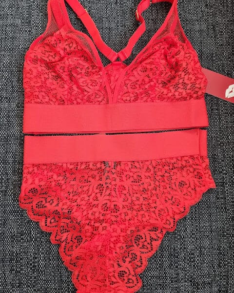 A red lace bodysuit with floral patterns is laid flat on a dark textured surface. The bodysuit features sheer mesh details, scalloped edges, adjustable straps, and a wide elastic waistband with a tag attached.