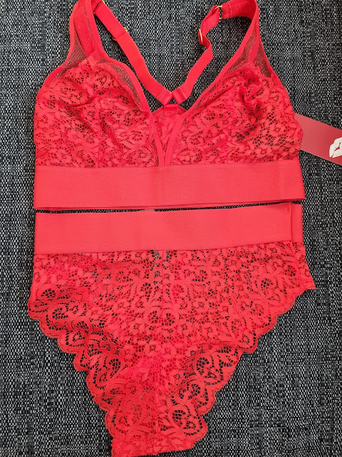 A red lace bodysuit with floral patterns is laid flat on a dark textured surface. The bodysuit features sheer mesh details, scalloped edges, adjustable straps, and a wide elastic waistband with a tag attached.