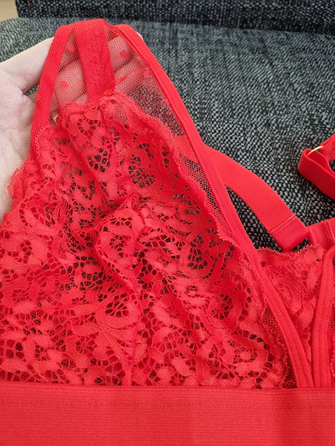 A close-up of a hand holding a red lace bralette with sheer mesh and floral lace details. The background features a textured dark grey fabric, possibly a sofa. A strap and part of the bralette's band are visible on the right side.
