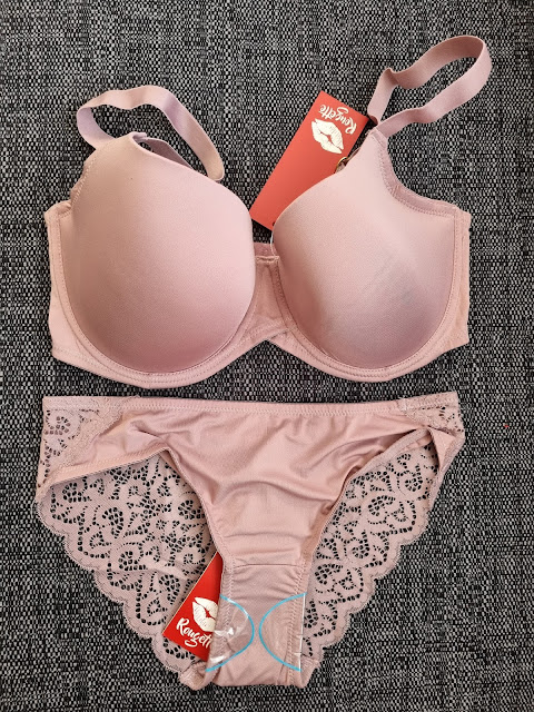 A blush-pink bra and matching knickers set is laid out on a dark textured surface. The bra has smooth, padded cups with adjustable straps, and the knickers feature a lace trim with a floral pattern. Both pieces have red tags with a lips graphic attached.