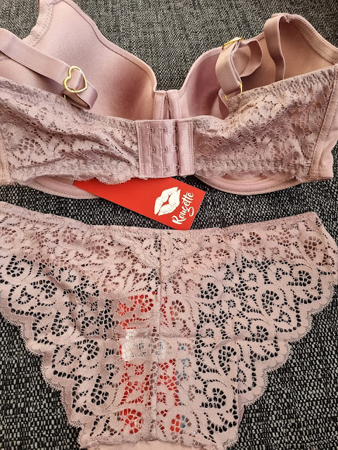 A mauve lace lingerie set is displayed on a dark textured fabric. The bra features heart-shaped metal accents and multiple hook fastenings, while the matching knickers have intricate floral lace patterns and scalloped edges. A red tag with a kiss mark is visible.