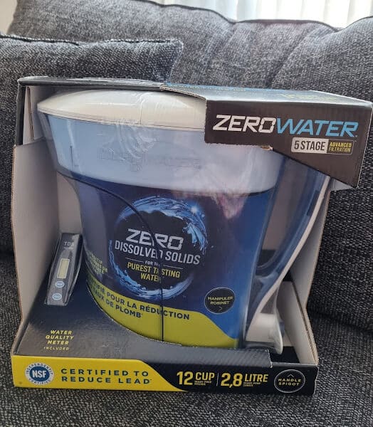 A ZeroWater 12-cup water filter jug is shown in its box, featuring 5-stage advanced filtration. The packaging highlights “Zero Dissolved Solids,” includes a total dissolved solids meter, and mentions certification to reduce lead. The jug is blue and white.