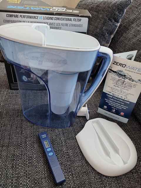 A blue ZeroWater filter jug with a white lid and filter is on a grey fabric surface. Nearby are a TDS water quality metre, a filter information card, and box packaging displaying product details and features.