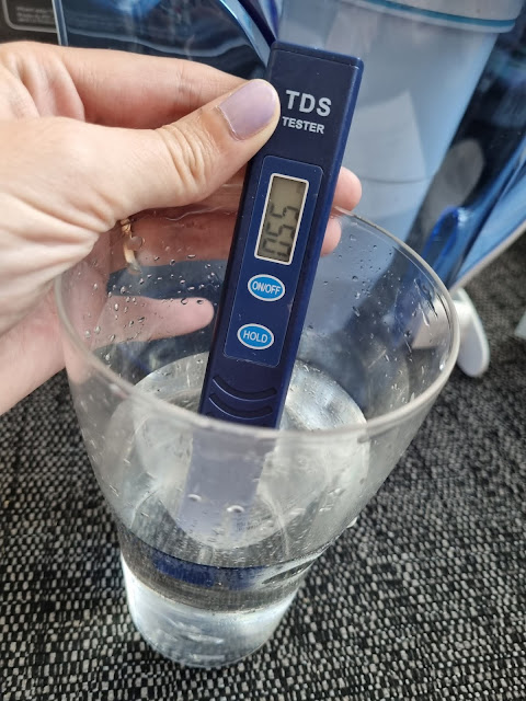 A hand holds a blue TDS tester with a digital display reading “163” in a glass of clear water. The glass sits on a dark textured surface, and a water filter jug is visible in the background. The tester has “ON/OFF” and “HOLD” buttons.