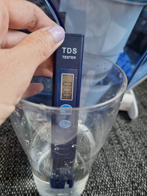 A hand with light purple nail varnish holds a blue TDS tester in a clear glass of water. The tester displays a reading of 009. The background shows a water filter jug on a dark textured surface.