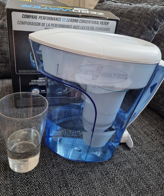 A blue ZeroWater filter jug sits on a grey sofa next to a clear drinking glass with some water. Behind them is the product box, featuring comparison information and branding. The jug features a white lid and filter visible inside.