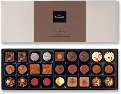 A rectangular box of assorted chocolates, each with unique shapes, toppings, and decorations, is arranged in five rows. The box lid is partially open, showing a label that reads “Everything Selection” from Hotel Chocolat.