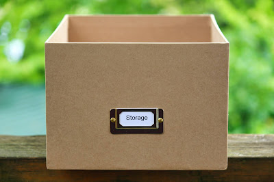 A plain, empty, brown cardboard box with a metal label holder on the front displaying a white label that reads "Storage." The box is placed on a wooden surface outdoors, with green foliage blurred in the background.