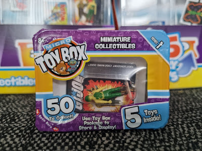 A close-up of a Micro Toy Box package featuring a tiny green toy car inside. The purple packaging highlights “Miniature Collectables,” “50 to collect!,” and “5 toys inside!” with cartoon graphics and a clear window showing the toy.