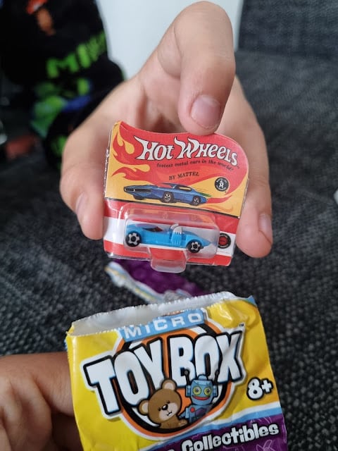 A hand holds a small Hot Wheels toy car still in packaging, with a bright red and yellow label. Another hand holds an opened packet labelled "Micro Toy Box Collectables 8+," showing part of a cartoon bear graphic on the wrapper.