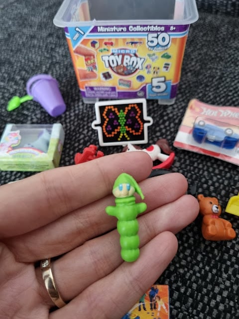A hand holds a small green caterpillar toy wearing a pointed cap. In the background are various miniature toys, including a bear, a purple bucket, a butterfly design, toy car, and a container labelled "Toy Box Miniature Collectables.
