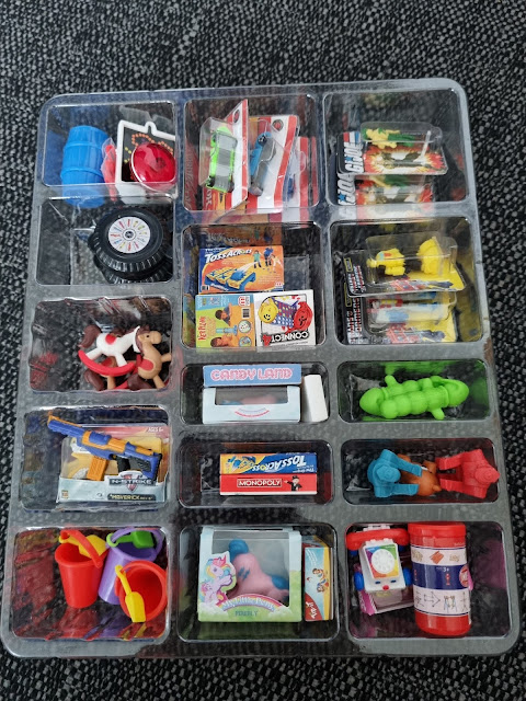 A plastic tray with sixteen compartments, each holding tiny, colourful toy miniatures resembling items like a tyre, water pistols, Hot Wheels cars, sweet boxes, a red bucket and spade set, a horse ride, bubbles, a caterpillar, and various small containers and figures.