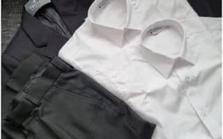 A neatly arranged dark suit with matching trousers, displayed on a hanger, lies next to two crisp, folded white dress shirts on a textured grey surface. The suit jacket and trousers are coordinated, and the shirts have classic pointed collars.