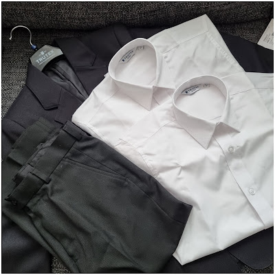 A neatly arranged dark suit with matching trousers, displayed on a hanger, lies next to two crisp, folded white dress shirts on a textured grey surface. The suit jacket and trousers are coordinated, and the shirts have classic pointed collars.