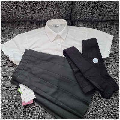 A neatly folded white short-sleeved collared shirt, a dark grey pleated skirt with tags attached, and a pair of folded black tights are displayed on a textured grey sofa.
