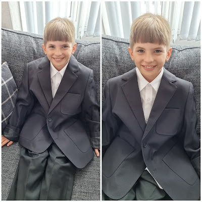 Side-by-side photos of a smiling young boy with light brown hair sitting on a grey sofa, wearing an oversized dark grey suit and white dress shirt. The suit jacket and trousers appear much too large for him. Light streams in through blinds behind him.
