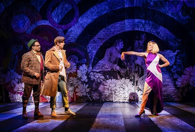 Three actors perform on stage. One wears a checked suit with a green hat, another wears a trench coat and cap, and the third, a woman in a purple dress with a white sash, stands with one arm extended dramatically. The backdrop is colourful with floral and artistic patterns.