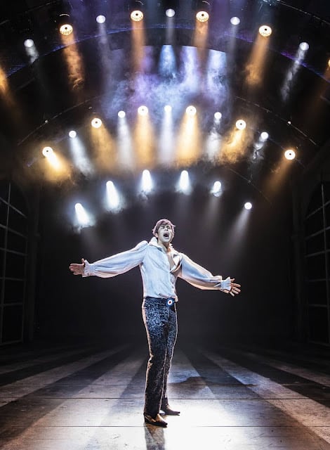 A performer in a white, flared shirt and patterned trousers stands centre stage under dramatic spotlights, arms outstretched and mouth open as if singing. The arched stage is illuminated with bright overhead lights and mist, creating a theatrical, energetic atmosphere.