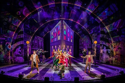 A vibrant stage scene shows actors in colourful costumes performing energetically under dramatic, purple lighting. A large, arched set with a house-like centrepiece features glowing stained glass windows, while musicians and cityscape props fill the background.