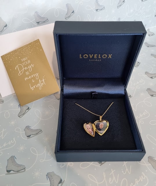 A gold heart-shaped locket necklace sits in a navy LOVELLOX London box. The locket shows two portrait photos inside. Next to the box is a gold greeting card that reads, "May your days be merry & bright." The background has a festive ice-skate pattern.