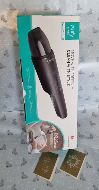 A Eufy HomeVac H11 handheld vacuum cleaner box stands upright on silver gift wrap with white reindeer and trees. The box is white and teal, showing the black vacuum cleaner. Two gold and green holiday cards lie on the lower right side.