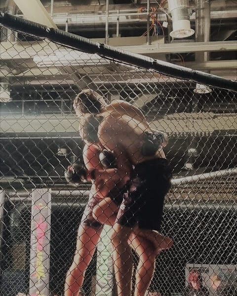 Two mixed martial artists grapple against the cage during a fight. One athlete lifts the other off the ground, pinning them to the chain-link fence inside the ring. Both wear gloves and shorts. Overhead industrial lights and pipes are visible above the cage.
