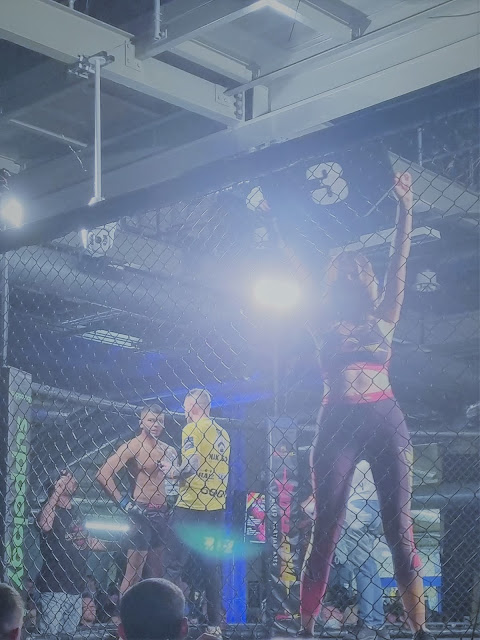 Inside a caged MMA ring, a woman in sportswear stands with arms raised by the fence, while a shirtless male fighter and a man in a yellow shirt stand in the background. Bright overhead lights illuminate the scene; spectators are visible outside the cage.