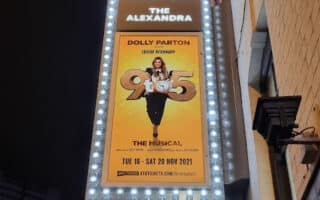 A lit theatre marquee for The Alexandra displays a poster for “9 to 5 The Musical” starring Dolly Parton and Louise Redknapp, with show dates from Tue 16 – Sat 20 Nov 2021, set against a yellow background and surrounded by bright bulbs.