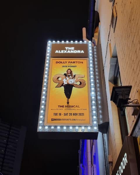 A lit theatre marquee for The Alexandra displays a poster for “9 to 5 The Musical” starring Dolly Parton and Louise Redknapp, with show dates from Tue 16 – Sat 20 Nov 2021, set against a yellow background and surrounded by bright bulbs.