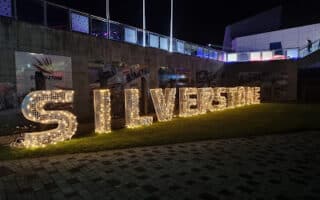Large illuminated letters spelling "SILVERSTONE" are displayed outdoors at night on a grassy area. Behind the letters are race-themed banners and a concrete wall. The scene is lit with warm lights, and some people are visible in the background.
