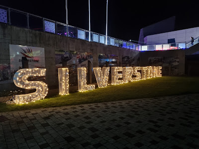 Large illuminated letters spelling "SILVERSTONE" are displayed outdoors at night on a grassy area. Behind the letters are race-themed banners and a concrete wall. The scene is lit with warm lights, and some people are visible in the background.
