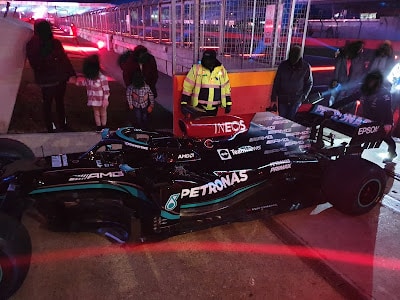 A black Formula 1 racing car with teal accents and "PETRONAS" branding is parked trackside at night. Several people, including a worker in a yellow jacket, stand nearby. Red track lights and barriers are visible in the background.