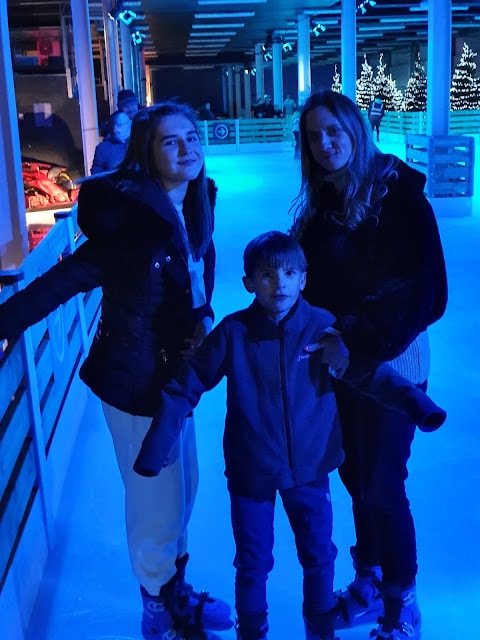 Three people—two women and a young boy—stand together on an ice rink under blue lighting. They wear winter clothes and ice skates. Behind them, festive lights and Christmas trees can be seen, creating a cheerful winter atmosphere.
