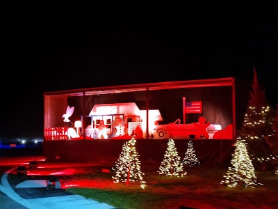 A night-time scene with a festive holiday display featuring red-lit cut-outs of a retro diner, a classic car, and people. Several small Christmas trees with white fairy lights are in the foreground, and the background is dark. The display is brightly illuminated.
