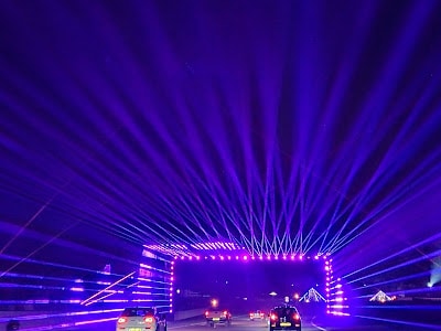Cars drive through a tunnel-like structure illuminated by bright purple and blue lights at night. Beams of light extend outward from the structure, creating a striking and colourful visual effect across the dark sky and roadway.