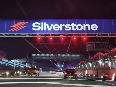 Night-time view of a car driving on a racetrack under a large illuminated sign that reads “Silverstone.” Red lights are overhead. The track is bordered by barriers and lit buildings, with additional lights and structures visible in the background.
