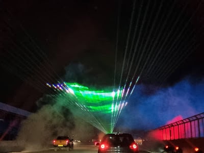 Cars drive on a road at night under a display of green and blue laser lights and smoke, creating colourful beams across the dark sky. The scene appears festive and dramatic, with illuminated smoke adding to the vibrant atmosphere.