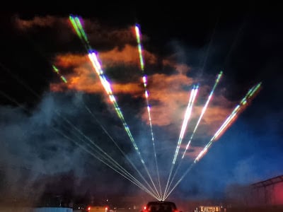 Colourful laser beams shoot upwards from a central point behind a car at night, piercing through clouds and mist. The beams fan out in different directions, creating rainbow streaks across the dark, smoky sky. Buildings and faint red lights are visible below.