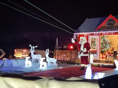 A house decorated with Christmas lights features a person dressed as Father Christmas waving beside glowing reindeer and sleigh figures. A lit Christmas tree is visible through the window. Two cars with headlights on are in the background.