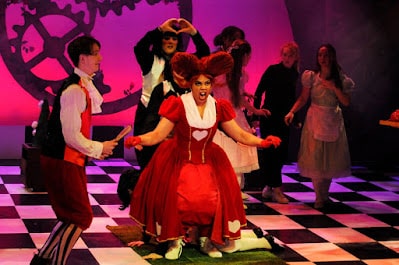 Actors perform on a chequered stage with a large gear backdrop. At centre, a woman in a red “Queen of Hearts” costume kneels, shouting. Other actors in whimsical costumes, including a rabbit and maids, surround her. The lighting is dramatic and colourful.