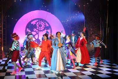 A vibrant stage scene shows actors in colourful, whimsical costumes dancing on a black-and-white chequered floor. A woman in a blue dress stands centre stage. Behind them, a large pink-lit backdrop features an ornate clock face design, with dramatic theatrical lighting.