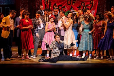 A lively stage scene shows a group of young people in colourful 1950s-style formal wear, gathered round a man in a leopard-print jacket sitting on the floor with arms wide. A red “Class of 1959” banner hangs behind them, evoking a school dance or prom setting.
