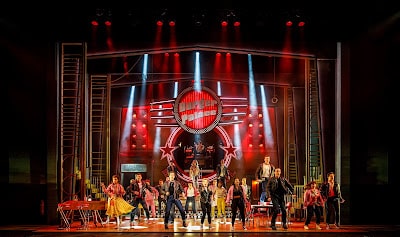A large musical ensemble performs on a colourful stage with vibrant red and yellow lighting. A woman stands atop a platform under a circular sign. Dancers in retro costumes move energetically below, framed by bright spotlights and theatrical set pieces.