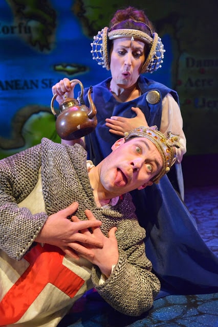 A man in chain mail with a crown and a red cross tunic clutches his chest and sticks out his tongue dramatically. Behind him, a woman in a blue mediaeval gown and elaborate headdress holds a teapot, appearing to pour onto his head, both with exaggerated expressions.