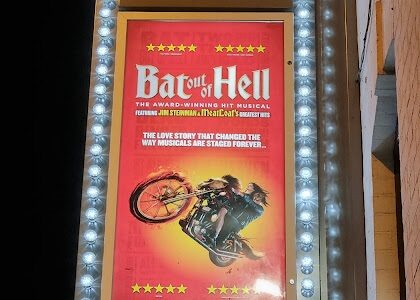 A lit theatre marquee reads “THE ALEXANDRA” above a poster for “Bat Out of Hell: The Musical.” The poster shows a man on a motorbike with flames, flying through the air, and promotional text in yellow and white on a red background.