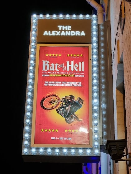 A lit theatre marquee reads “THE ALEXANDRA” above a poster for “Bat Out of Hell: The Musical.” The poster shows a man on a motorbike with flames, flying through the air, and promotional text in yellow and white on a red background.